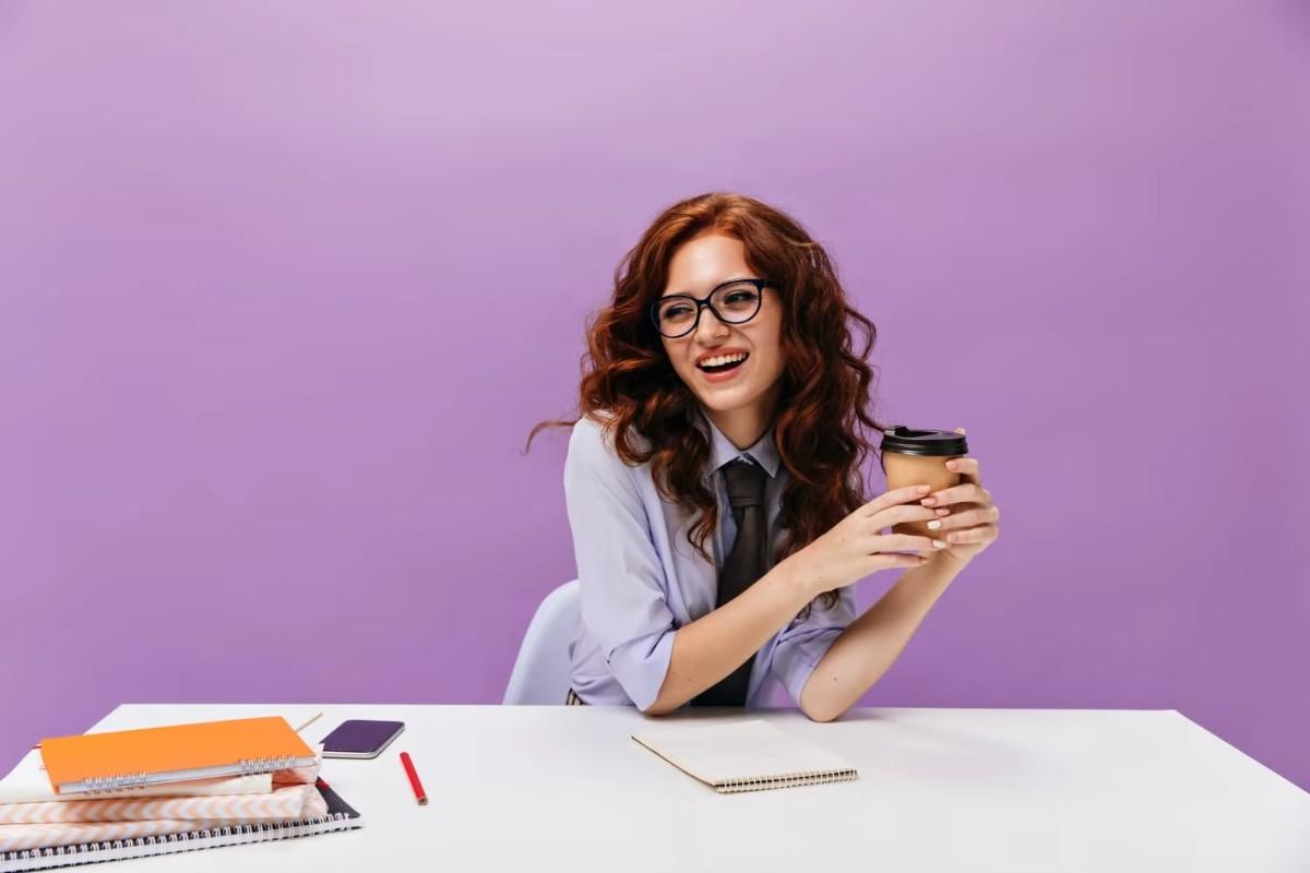 A stylish young woman wearing glasses, sporting a radiant smile, and sipping from her cup.